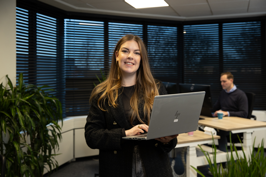 Vrouwelijke medewerker staat met laptop in haar handen, op de achtergrond zie je een collega aan een bureau.